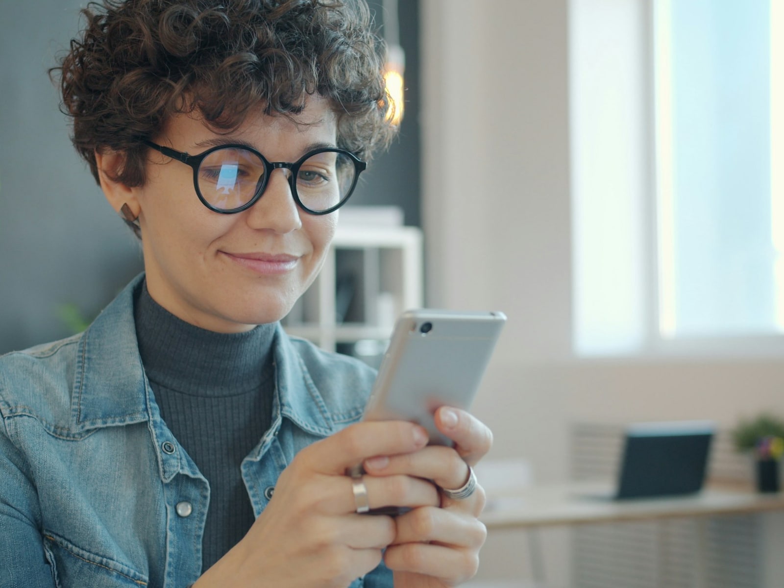 Pessoa de óculos sorrindo ao olhar o celular em um escritório claro
