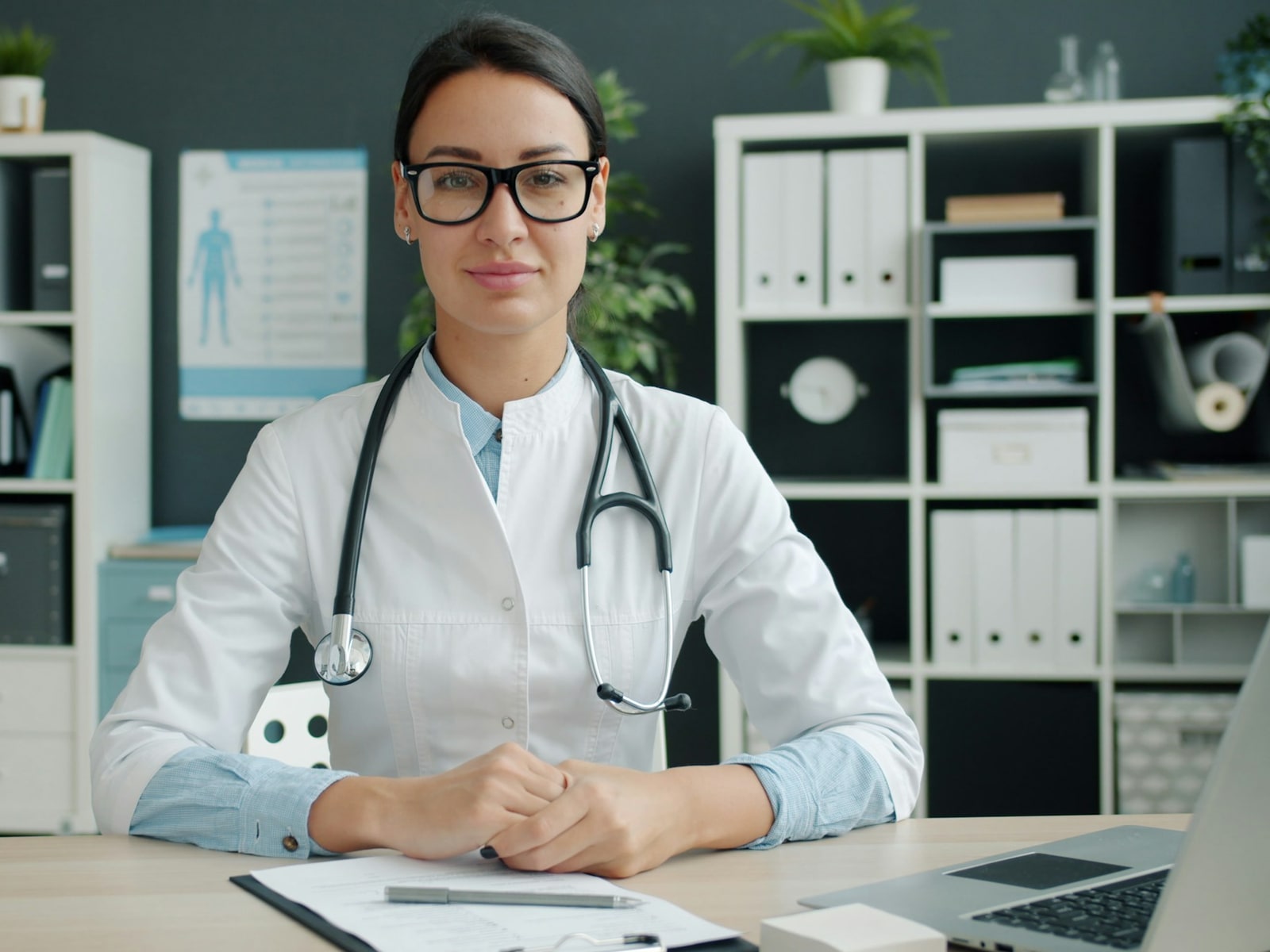 Médica de jaleco e estetoscópio sentada à mesa do consultório, com prancheta e computador
